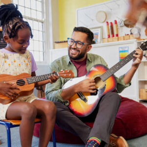 Happy male teacher assisting an african kid to play musical instrument in classroom at school. Happy teacher playing acoustic guitar and singing while having music class with girl. Cute little girl learn to play ukulele with male teacher.